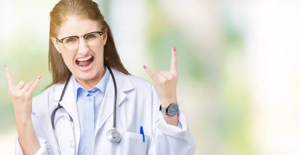 Middle age mature doctor woman wearing medical coat over isolated background shouting with crazy expression doing rock symbol with hands up. Music star. Heavy concept.