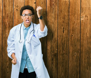 Genç Afro-Amerikan doktor kadın öfkeli ve sinirli sinirli ve öfke ile bağırarak süre öfkeli yumruk yükselterek izole arka plan üzerinde tıbbi bir palto giyiyor. Öfke ve agresif kavramı.