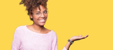 Güzel genç Afro-Amerikan kadın neşeli sunulması ve kameraya bakarak avucunun ile işaret gülümseyen izole arka plan üzerinde gözlük.