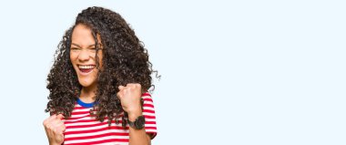 Kıvırcık saçlı genç güzel kadın çizgili t-shirt giyen çok mutlu ve heyecanlı kolları kaldırdı ile kazanan jest yapıyor, gülümseyerek ve başarı için çığlık. Kutlama konsepti.