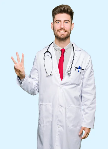 年轻英俊的医生男子穿着医疗外套显示和手指第三 而微笑着自信和快乐 — 图库照片
