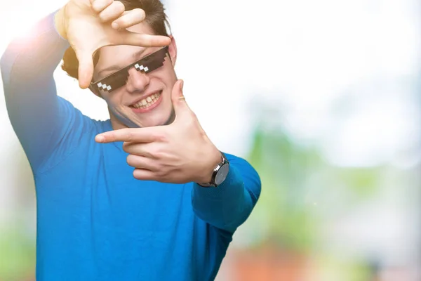 Young man wearing funny thug life glasses over isolated background smiling making frame with hands and fingers with happy face. Creativity and photography concept.