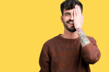 El yüz ve sürpriz duygu kendinden emin gülümseme ile tek gözüyle kapsayan izole arka plan üzerinde kış kazak giyen genç yakışıklı adam.