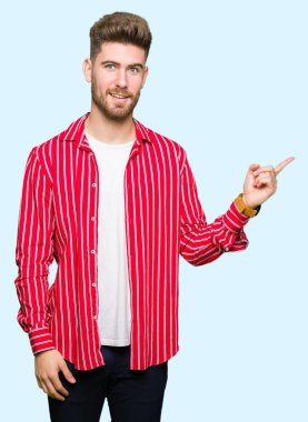 Kameraya bakarak yan el ve parmak ile işaret, yüzünde kocaman bir gülümseme ile kırmızı gömlek giymiş genç yakışıklı adam.