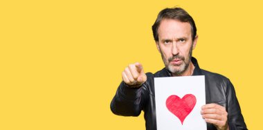 Yakışıklı orta yaş erkek holding kağıt kırmızı kalp kamera ve size parmak ile işaret ile el işareti, pozitif ve kendinden emin bir jest ön