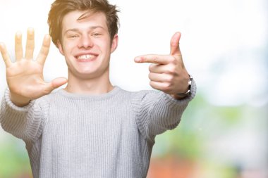 Gösterilen arka plan üzerinde kış kazak giyen genç yakışıklı adam izole ve parmakları ile işaret kendinden emin ve mutlu gülümseyerek süre yedi numara.