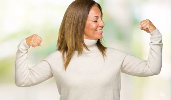 Beautiful middle age adult woman wearing winter sweater over isolated background showing arms muscles smiling proud. Fitness concept.