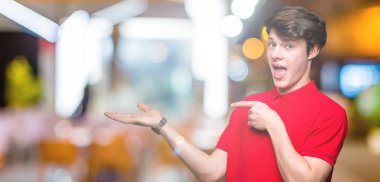 Hayret ve el ile sunulması ve parmak ile işaret ederken kameraya gülümseyen izole arka plan üzerinde kırmızı t-shirt giyen genç yakışıklı adam.