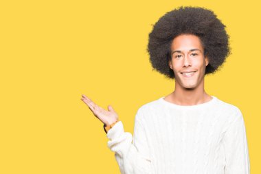 Neşeli sunulması ve kameraya bakarak avucunun ile işaret gülümseyen kış kazak giyen afro saçlı genç Afro-Amerikan adam.