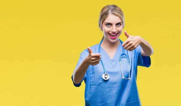 年轻美丽的金发医生护士护士妇女在孤立的背景下批准做积极的手势与手 竖起大拇指微笑 并为成功高兴 看着镜头 赢家的手势 — 图库照片