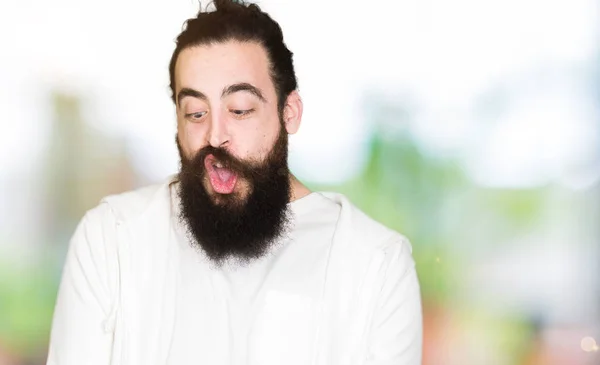 Young man with long hair and beard wearing sporty sweatshirt making fish face with lips, crazy and comical gesture. Funny expression.