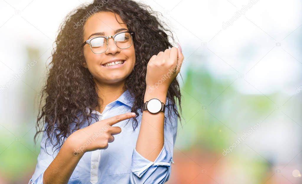 Young beautiful business girl with curly hair wearing glasses In hurry pointing to watch time, impatience, upset and angry for deadline delay