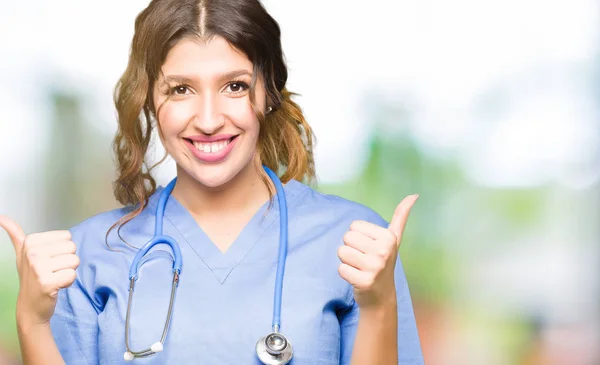 年轻的成年医生妇女穿着医疗制服的成功标志做积极的姿态与手 竖起大拇指微笑和快乐 看着相机欢快的表情 胜利者的手势 — 图库照片