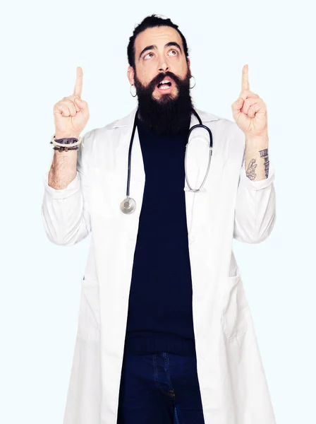 聴診器と医療のコートを身に着けている長い髪の医者はびっくりし 驚き見て 指で指して 腕を上げた — ストック写真