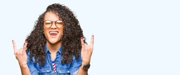 メガネをかけた巻き毛の若い美しい女性が 手を上げてロックシンボルをやっているクレイジーな表情で叫ぶ 音楽スターだヘビーコンセプト — ストック写真