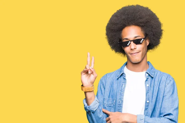 Young african american man with afro hair wearing thug life glasses smiling with happy face winking at the camera doing victory sign. Number two.