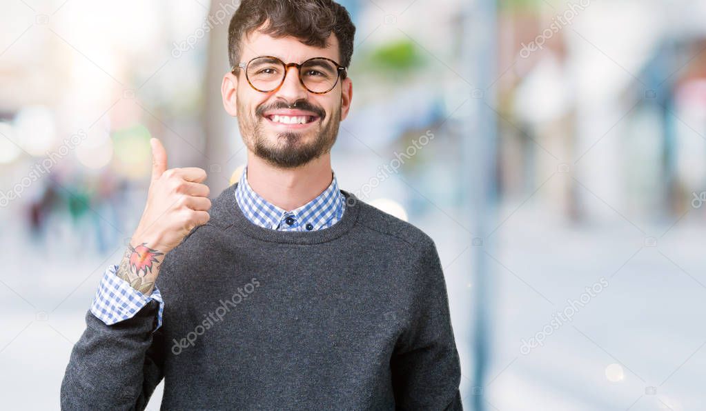 Young handsome smart man wearing glasses over isolated background doing happy thumbs up gesture with hand. Approving expression looking at the camera showing success.