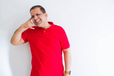 Beyaz duvarın üzerinde kırmızı tişört giyen orta yaşlı adam telefonda konuşmak gibi el ve parmaklarla telefon hareketi yaparak gülümsüyor. İletişim kavramları.