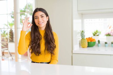 Genç güzel kadın evde beyaz masada gülümseyerek olumlu el ve parmakları ile ok işareti yapıyor. Başarılı ifade.