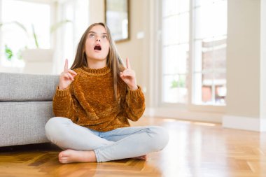 Evde yerde oturan güzel genç kız çocuk şaşırır ve görünümlü ve parmaklar ve yükseltilmiş kollar ile işaret şaşırttı.