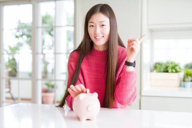 Güzel Asyalı kadın piggy çanta içinde bir sikke koyarak çok mutlu yan el ve parmak ile işaret