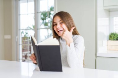 Güzel genç kız evde büyük bir gülümseme ok işareti yapıyor mutlu bir kitap okuma, parmakları ile başparmak, mükemmel işareti