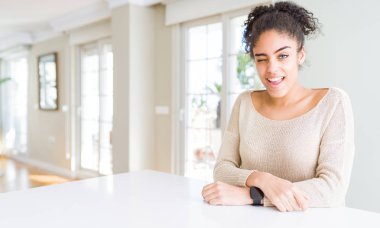 Afro saçlı güzel genç afro-amerikan kadın evde masada oturan seksi ifade, neşeli ve mutlu yüz ile kameraya bakarak göz kırpıyor.