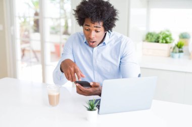 African American business man using smartphone and laptop scared in shock with a surprise face, afraid and excited with fear expression