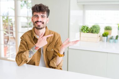 Sıradan bir ceket giyen genç adam beyaz masaya oturmuş kameraya hayret ediyor ve gülümsüyor..