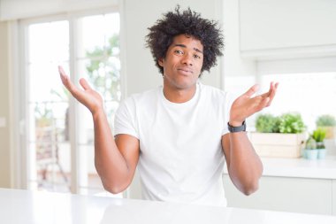 Sıradan beyaz tişörtler giyen Afro-Amerikan genç adam evinde oturuyor ve elleri ve kolları havada, kafası karışık bir ifade takınıyor. Şüphe kavramı.