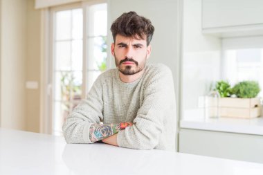 Genç adam beyaz masa şüpheci ve sinir, onaylamayan ifade yüz çapraz kolları üzerinde oturan rahat kazak giyiyor. Negatif kişi.