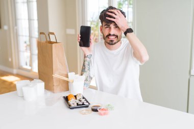 Ev teslimatından Asya suşisi yiyen ve akıllı telefon ekranı gösteren genç bir adam elleri kafasında, utanç ve şaşkınlık dolu, sinirli ve hayal kırıklığına uğramış bir halde. Hata için korku ve üzüntü.