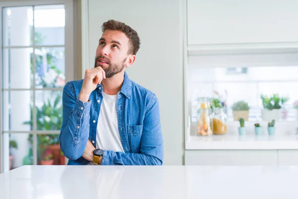 Handsome man at home with hand on chin thinking about question, pensive expression. Smiling with thoughtful face. Doubt concept.