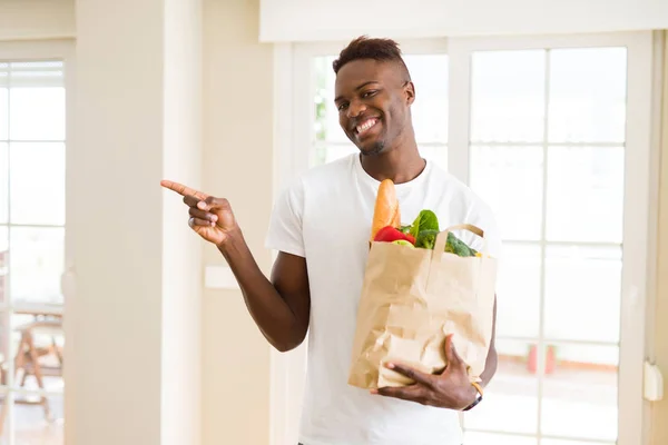 African american man holding paper bag full of fresh groceries very happy pointing with hand and finger to the side