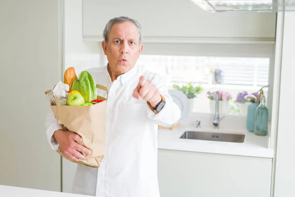 Handsome senior man holding a paper bag of fresh groceries at the kitchen pointing with finger to the camera and to you, hand sign, positive and confident gesture from the front