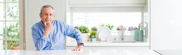 Wide angle perspective of handsome senior man at home looking confident at the camera with smile with crossed arms and hand raised on chin. Thinking positive.