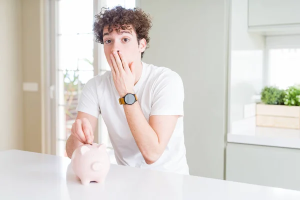 Young man investing money using piggy bank at home cover mouth with hand shocked with shame for mistake, expression of fear, scared in silence, secret concept