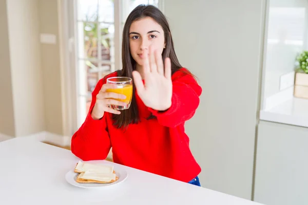 Beautiful young woman eating toasts and orange juice for snack or breakfast with open hand doing stop sign with serious and confident expression, defense gesture