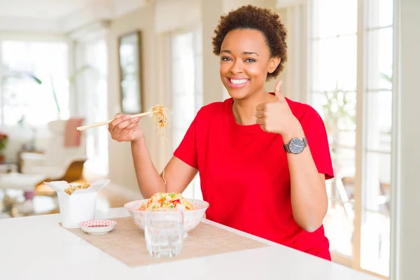 Young african american woman with afro hair eating asian food at home happy with big smile doing ok sign, thumb up with fingers, excellent sign