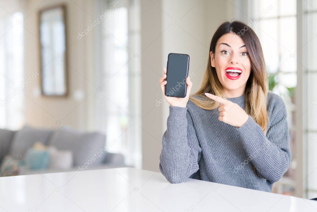 Young beautiful woman showing smartphone screen at home very happy pointing with hand and finger