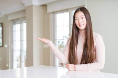 Güzel Asyalı kadın beyaz masada rahat kazak giyen neşeli sunan gülümseyerek ve kameraya bakarak avuç içi ile işaret.