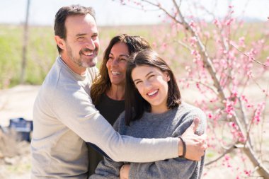 Üç gülümseyen güzel ailesi neşeli ve sarılma şeftali bahçesinde bahar güneşli gün zevk pembe yaprakları ile