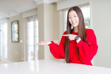 Beyaz masada kırmızı kazak giyen güzel Asyalı kadın şaşırttı ve el ile sunarken kameraya gülümseyerek ve parmak ile işaret.