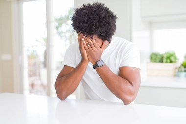 Beyaz günlük tişörtler giyen genç Afro-Amerikalı adam evde oturup ağlarken suratını ellerle kapatıyor. Depresyon kavramı.