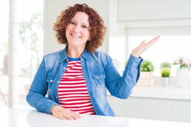 Evde kot ceket giyen kıvırcık saçlı orta yaş yaşlı kadın neşeli sunum gülümseyerek ve kameraya bakarak avuç içi ile işaret.