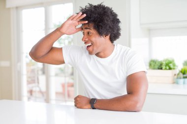 Beyaz günlük tişörtler giyen genç Afro-Amerikan bir adam evinde mutlu bir şekilde oturuyor ve gülümsüyor. Konsept aranıyor.