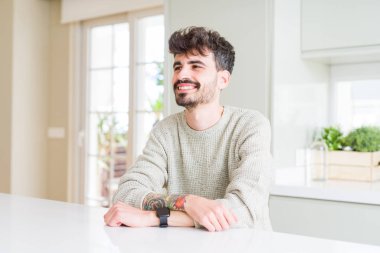 Gündelik kazak giyen genç bir adam beyaz masada oturuyor. Yüzünde gülümseme, doğal bir ifade ile yan yana bakıyor. Kendinden emin gülen.