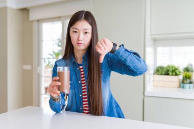 Güzel Asyalı kadın kızgın yüzü ile çikolata milkshake taze bir bardak içme, aşağı başparmak ile sevmediğim gösteren negatif işareti, ret kavramı
