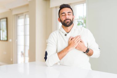 Yakışıklı İspanyol adam evde rahat beyaz kazak giyen kapalı gözleri ve yüzünde minnettar jest ile göğüs elleri ile gülümseyerek. Sağlık kavramı.