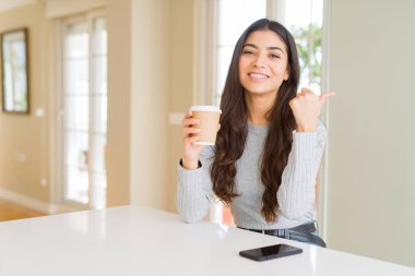 Genç kadın evde bir fincan kahve içme işaret ve mutlu yüz gülümseyen yanında başparmak ile gösteren